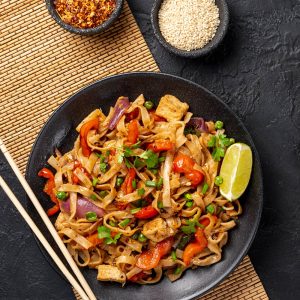 pad thai with chopsticks in a black bowl on a bamboo mat on black background