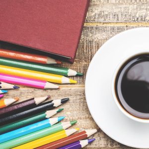 Cup of coffee on a white saucer with colored pencils and a maroon book on a wooden table.