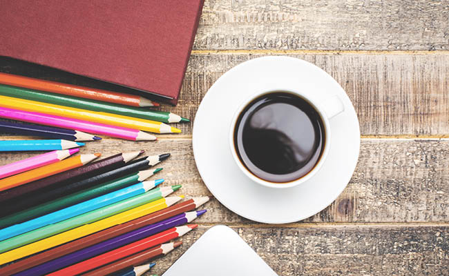 Cup of coffee on a white saucer with colored pencils and a maroon book on a wooden table.