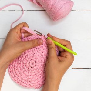 Hands crocheting a pink yarn circle with a green crochet hook on a white wooden table.