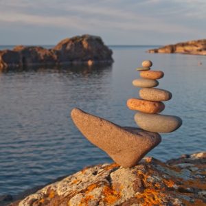 A stack of balanced stones on a lakeside rock at sunset, with calm water and distant rocky landforms in the background.