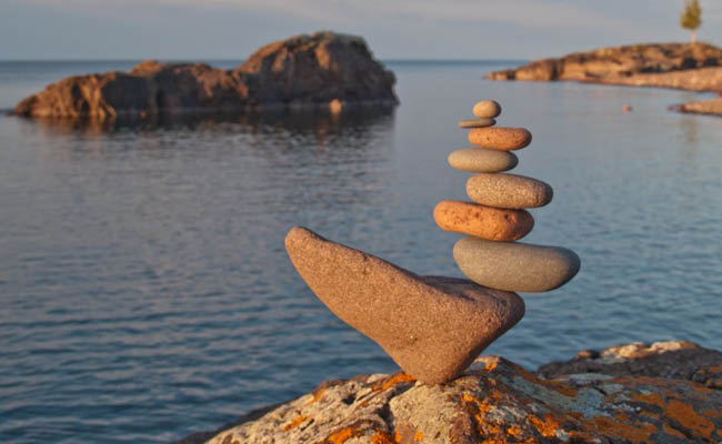 A stack of balanced stones on a lakeside rock at sunset, with calm water and distant rocky landforms in the background.
